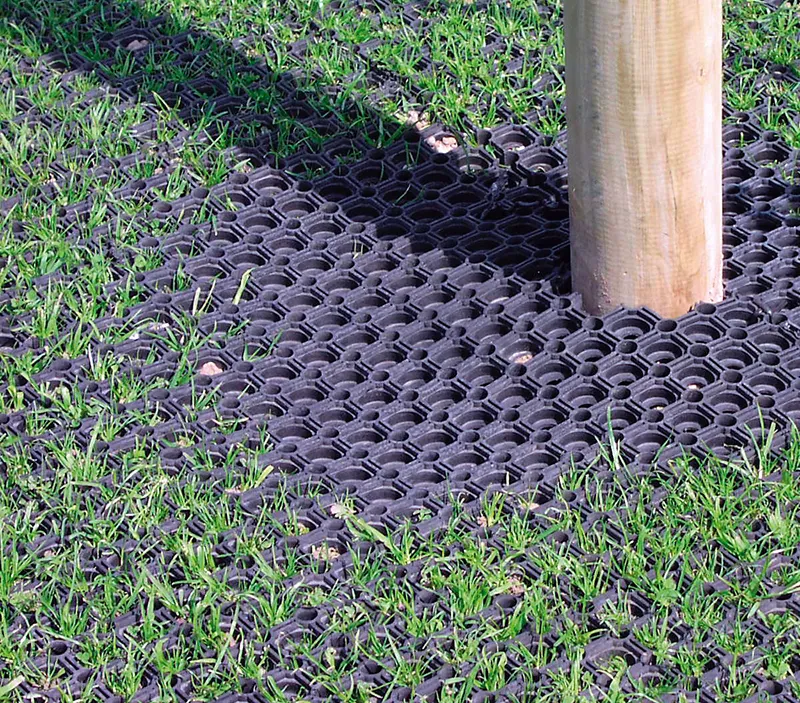A close-up view of green grass growing through a black rubber mat with circular holes, placed around the base of a wooden post for ground protection.