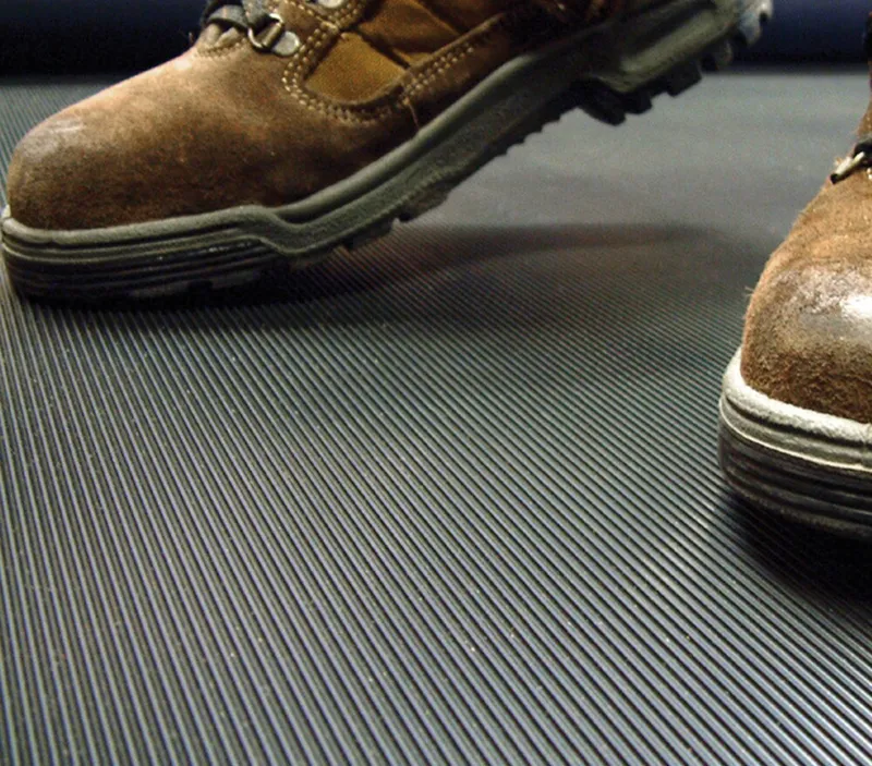 Close-up of two brown suede work boots standing on a black industrial anti-slip rubber mat with fine ridges.