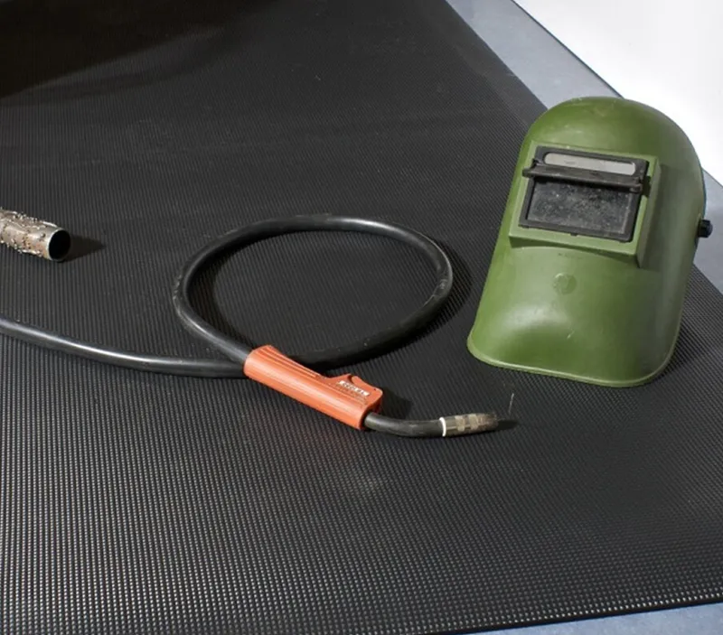 A green welding helmet and a welding torch with a black cable rest on a textured black mat.