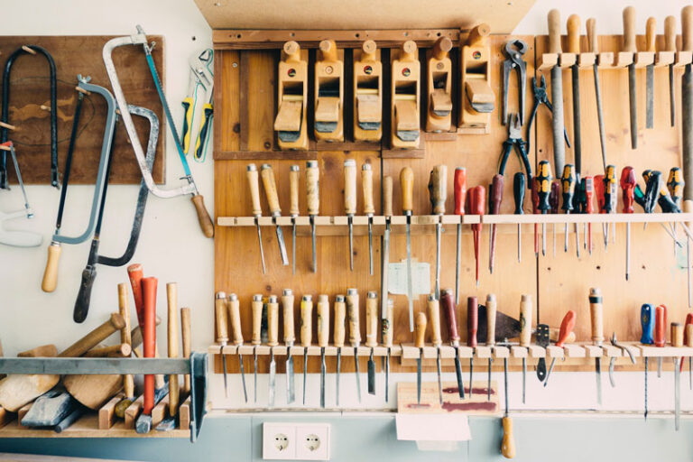 A neatly organised workshop wall with various woodworking tools, including saws, hammers, hand planes, chisels, and screwdrivers, all arranged on wooden racks and hooks above a workbench.
