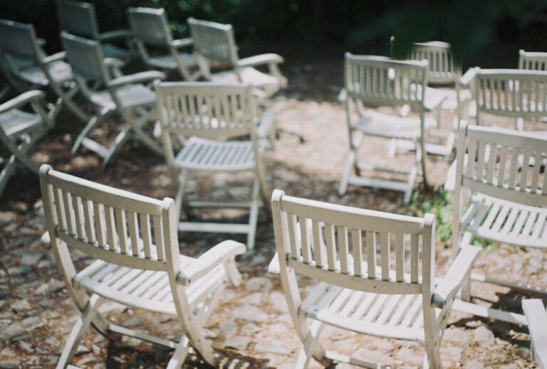 A group of empty white wooden chairs arranged outdoors on a stone surface, with sunlight filtering through trees, creating a peaceful and quiet setting.