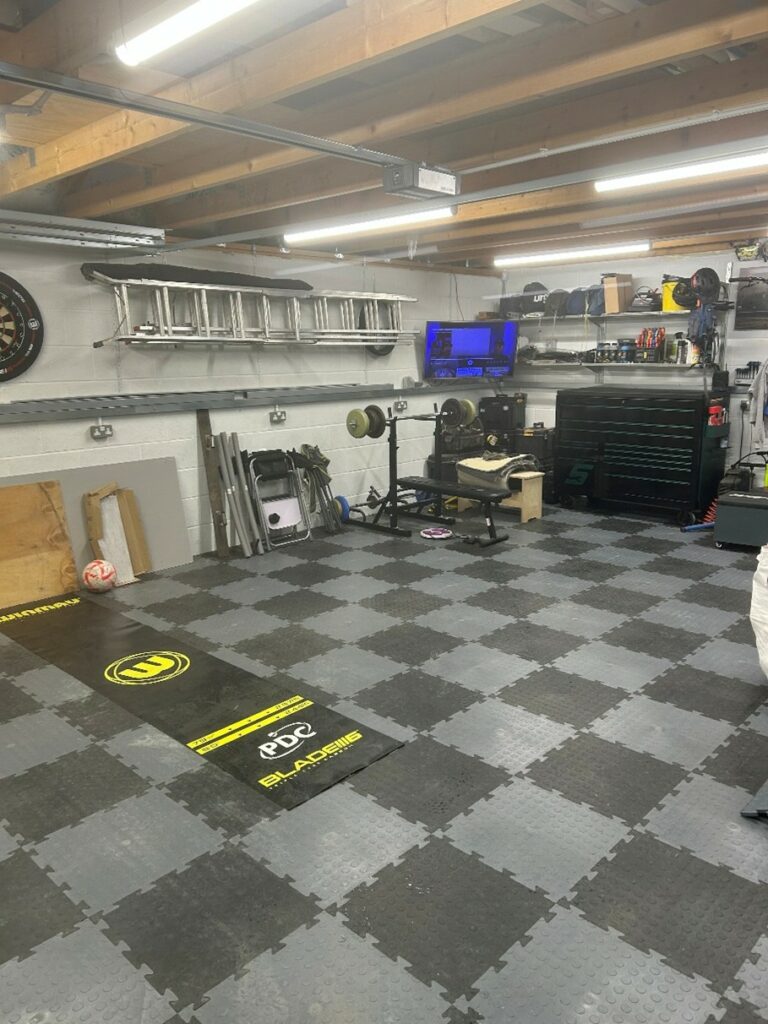 A clean garage features gym equipment, a dartboard, ladders mounted on the wall, a TV, organised tools, a workbench, and interlocking foam floor tiles. A football and mats are visible on the floor.